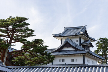 Upper view of Kanazawa Castle tower in Japan with traditional tiled roofs and decorative gables framed by pine trees under soft daylight and clear sky