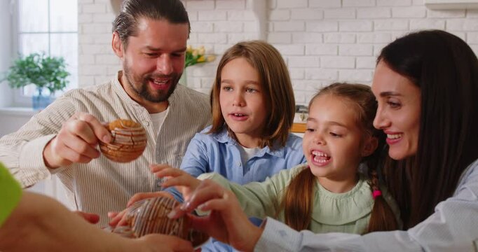 Cheerful family enjoying eating tasty pastries for breakfast, posing in kitchen, smelling appetizing buns, applauding and smiling. Parents and their children visiting guests, sitting at dinner table