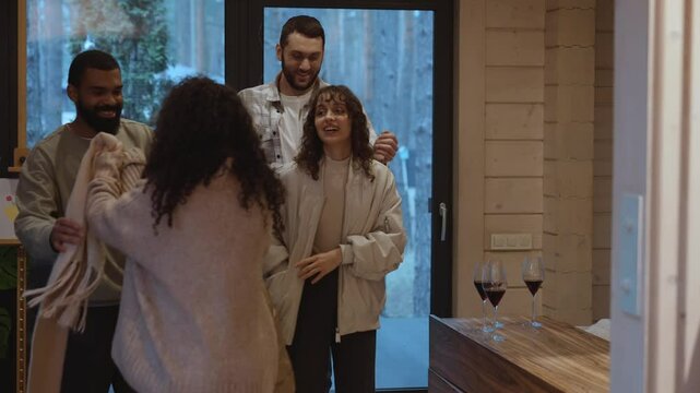 Entering the living room in the evening, a Black man in his early 20s helps his Black adult woman take off her jacket while a European 20-year-old couple undress nearby.