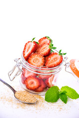 Glass jar with sliced ​​strawberries and brown sugar on white background. Fresh berries with mint and cane sugar. Summer dessert preparation.