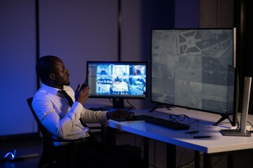 African American security guard looking at desktop computer screen while speaking on walkie talkie