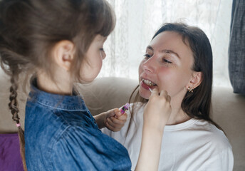 Daughter puts makeup on her mother face and plays around. Caring parent allows her child to paint her face. Happy childhood. Concept of loving family and fun. little girl doing her makeup