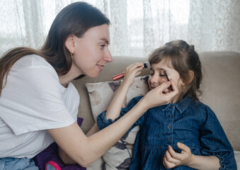 Mother putting makeup on her daughter face and they fool around together. Communication between women in the family. Fun time. Happy family concept. little girl doing her makeup