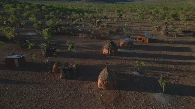 Drone flies over a Himba tribe camp in Namibia during golden hour, revealing red earth, traditional huts, scattered trees, and distant mountains in stunning light.