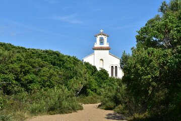 Chapelle blanche cachée derrière la végétation au bout d'un chemin en sable.