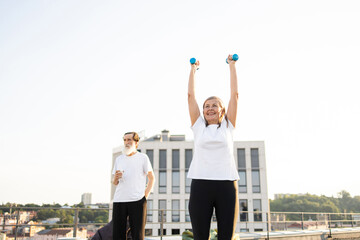 Fototapeta premium Elderly couple exercising together outdoors in urban area, woman lifting dumbbells, man observing. Healthy lifestyle, fitness, staying active in middle age.
