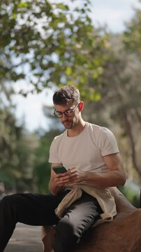 A young man sits on a log in a park, smiling as he uses his phone. He's wearing a white t-shirt, dark pants, glasses, and has a beige overshirt tied around his waist.