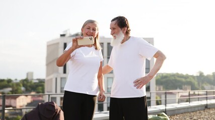 Elderly man and woman smiling while taking selfie outdoors, showing thumbs up, promoting active lifestyle and positivity in golden years.