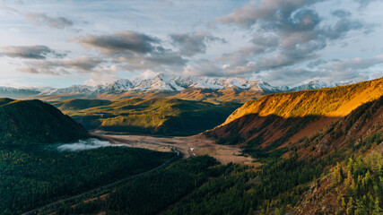 Golden sunlight illuminates autumnal slopes of mountains and valley with coniferous forest, river and fog under cloudy sky in altai, russia