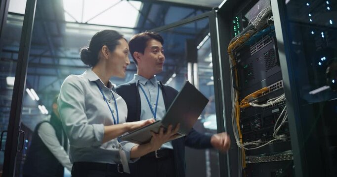 Male Network Engineer and Asian Female Data Analyst Collaborate on a Laptop Computer in a Modern Data Center Facility, Optimizing Network Performance and Data Flow Across Servers