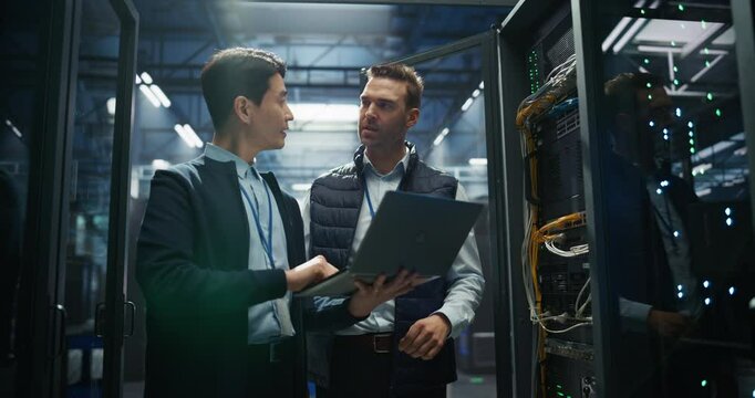 Asian IT Technician with a Laptop Computer and Caucasian Male Engineer Colleague are Talking in Data Center while Working Next to Server Racks. Running Diagnostics and Doing Maintenance Work