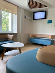 Interior Of A Modern And Bright Clinic Or Hospital Waiting Area With Light Blue Sofas, A White Coffee Table, Window, And Wall-Mounted TV. Healthcare Facility Design.