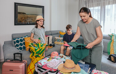 Family packing suitcases for summer holidays in living room. Smiling daughter holding inflatable pineapple float and mother folding swimwear to suitcase trip while son playing with ping pong paddles.