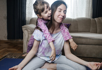 Young mother tries to meditate and do a home workout while her playful daughter gets in her way. Interrupted yoga at home and parenting reality. Mom and daughter having fun