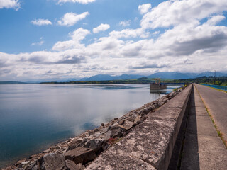 A paved road on the dam of Liptovská Mara reservoir in Slovakia, with the waterworks and High Tatras mountains visible under a partly cloudy sky.
