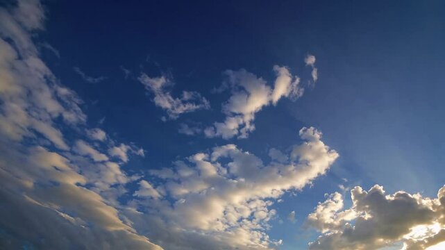 blue sky with scattered clouds at sunset. The clouds are mostly white and fluffy, with some darker areas towards the bottom of the frame. The sun is setting, casting a warm glow on the clouds.