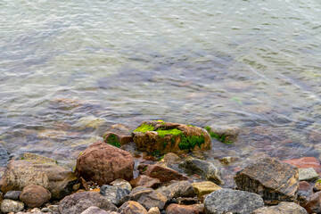 Rocky shoreline with colorful stones partially submerged in clear water. Some rocks are covered with green moss and algae. A natural scene from a seaside or lakeside environment.