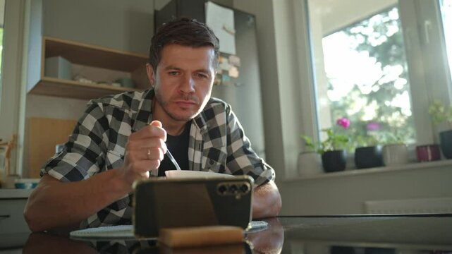 Man in plaid shirt eating oatmeal porridge from white bowl while using smartphone. Male looking at phone during breakfast. Concept of social media addiction, distraction, and digital dependency
