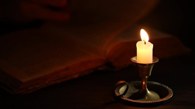 Page of an old book being turned over by a man hand with burning wax candle in bronze candlestick on wooden table in dark room at night, close up. Leisure reading books. Hand turning pages of a book