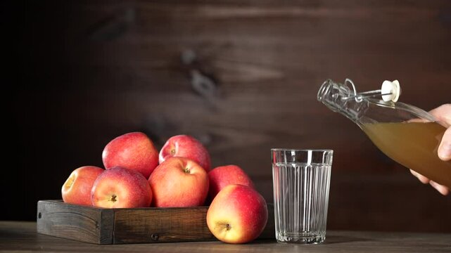 Man pouring apple cider from a glass bottle into a glass next to fresh red apples in wooden tray on kitchen table, close up