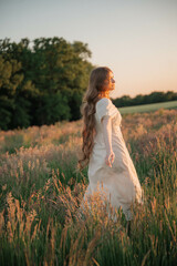 Brunette woman with long hair in a beige dress standing in a field at sunset, close-up portrait...