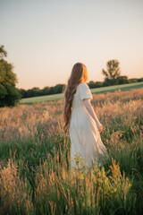 Brunette woman with long hair in a beige dress standing in a field at sunset, close-up portrait...
