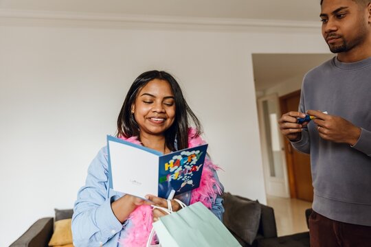 A young black woman wearing a pink feather boa reads a colorful birthday card while holding a gift bag indoors. A young man stands nearby, preparing to open a snack.
