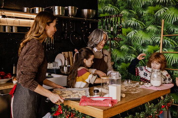 White school-aged children help their White adult mother and White grandmother cut shapes out of dough for future cookies at home in the kitchen. A concept of teamwork, bonding, and family routine.