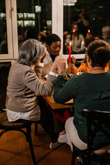 A Black adult man is sitting at a dinner table, holding hands with a white-skinned girl and two white-skinned women, and praying with closed eyes in a living room with floor-to-ceiling windows