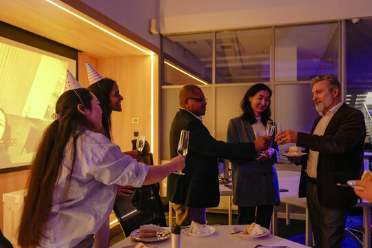 A senior White man in a brown blazer is clinking champagne glasses while holding a cake and celebrating his retirement with a multinational group of colleagues wearing party hats in an office lounge. - Powered by Adobe