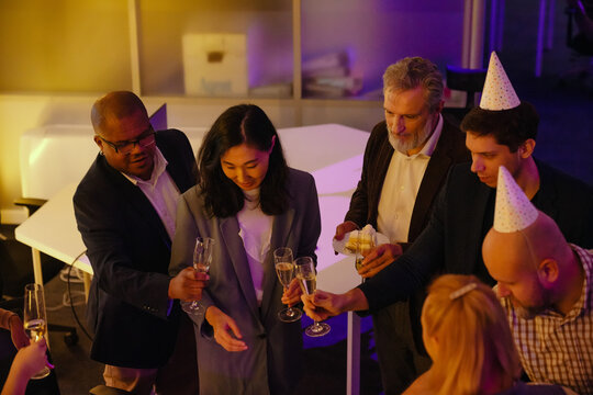 A senior White man in a brown blazer is holding a dessert plate while celebrating his retirement with a multinational group of colleagues in party hats with champagne glasses in an office at night.