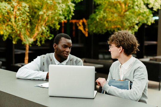 A young White man and an adult Black male mentor sitting at a long modern desk indoors, working on a laptop together during an internship program, both dressed in business casual sweaters and shirts.