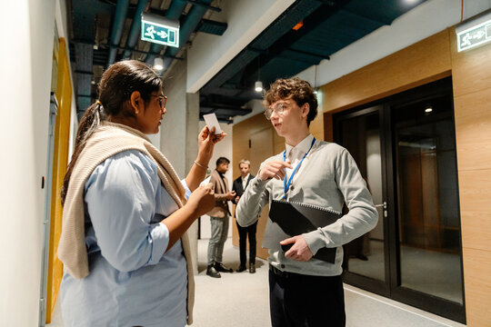 A young White man and a young Indian woman standing and discussing documents inside a hallway, both wearing business clothes, holding papers and notebooks, and having a conversation during work.