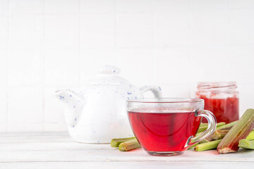 Organic natural rhubarb tea, sweet herbal plant based pink and green rhubarb hot tea drink on teapot and glass cup, on white wooden table with fresh rhubarb copy space