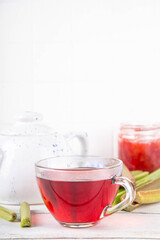 Organic natural rhubarb tea, sweet herbal plant based pink and green rhubarb hot tea drink on teapot and glass cup, on white wooden table with fresh rhubarb copy space