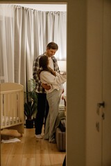 A young White pregnant woman standing beside her husband in a warmly lit bedroom nursery, wearing...