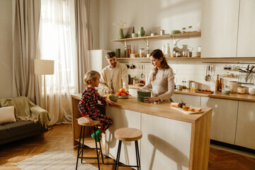 A school-age White boy in colorful pajamas sitting at a kitchen island with his White father in a cream cardigan and a White mother preparing breakfast in a sunlit modern kitchen with wooden accents.