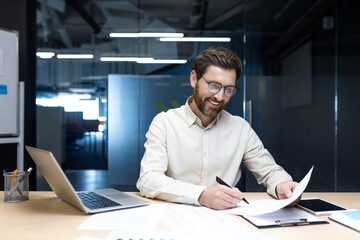 A successful and smiling young businessman sits in the office at a table with a laptop and signs...
