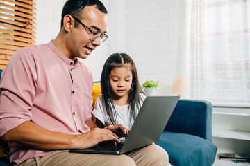 Smiling Asian father balances work with family time he works on his laptop while his daughter is engrossed in e-learning on computer. In their modern living room they create a moment of togetherness.