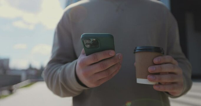 Close up of a man drinking to go coffee and browsing smartphone social networking at urban modern street. People and communication concept.