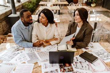 A young Black man and a Black woman meet with a female wedding planner. They sit at a table with wedding brochures and a laptop, discussing plans at a decorated venue.
