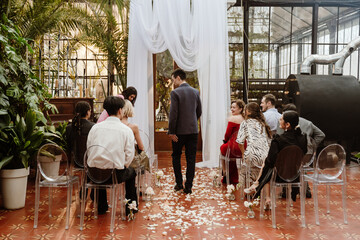 A group of wedding guests sits indoors in a hall with flower petals, transparent chairs, and a...