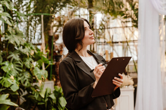 A young White female wedding planner holding a black clipboard, standing indoors in a greenhouse-style venue with lush green plants, wearing a black suit and white top during event preparation.