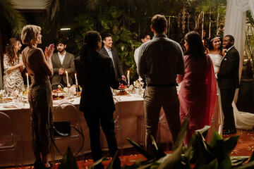 A Black bride and a Black groom are standing close to each other near a wedding arch while their international guests are standing around a table, looking at them and cheering