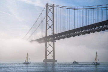 25 de Abril bridge in morning fog with sailboats on Tagus river