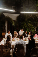 A young Black groom in a black tuxedo sitting beside his young Black bride in an off-shoulder white dress surrounded by a multinational group of wedding guests dining at an evening wedding reception.