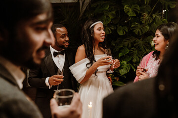 A Black bride in a wedding dress is holding a glass of champagne and talking with a smiling Latino woman while her groom is standing behind her with a glass and looking at her friend in a crowd