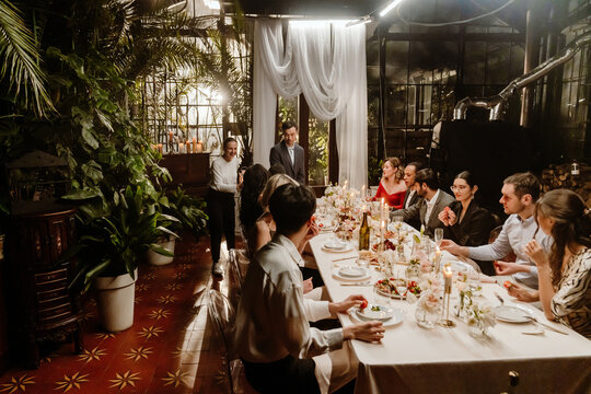 A group of adult guests seated at a long candlelit table enjoy an elegant wedding reception dinner indoors, surrounded by floral arrangements, tropical plants, and candles.