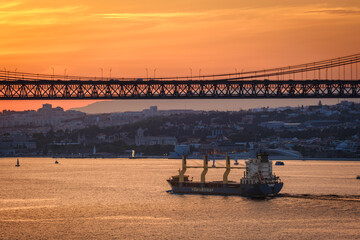Cargo ship passing under 25 de Abril bridge at colorful sunset, Lisbon