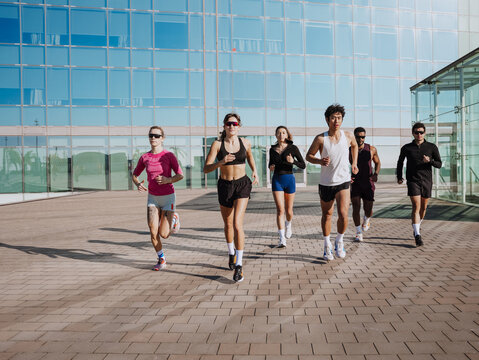 Seven young athletes run outdoors in front of a modern glass building. They wear athletic outfits and sunglasses and jog together on a sunny day.